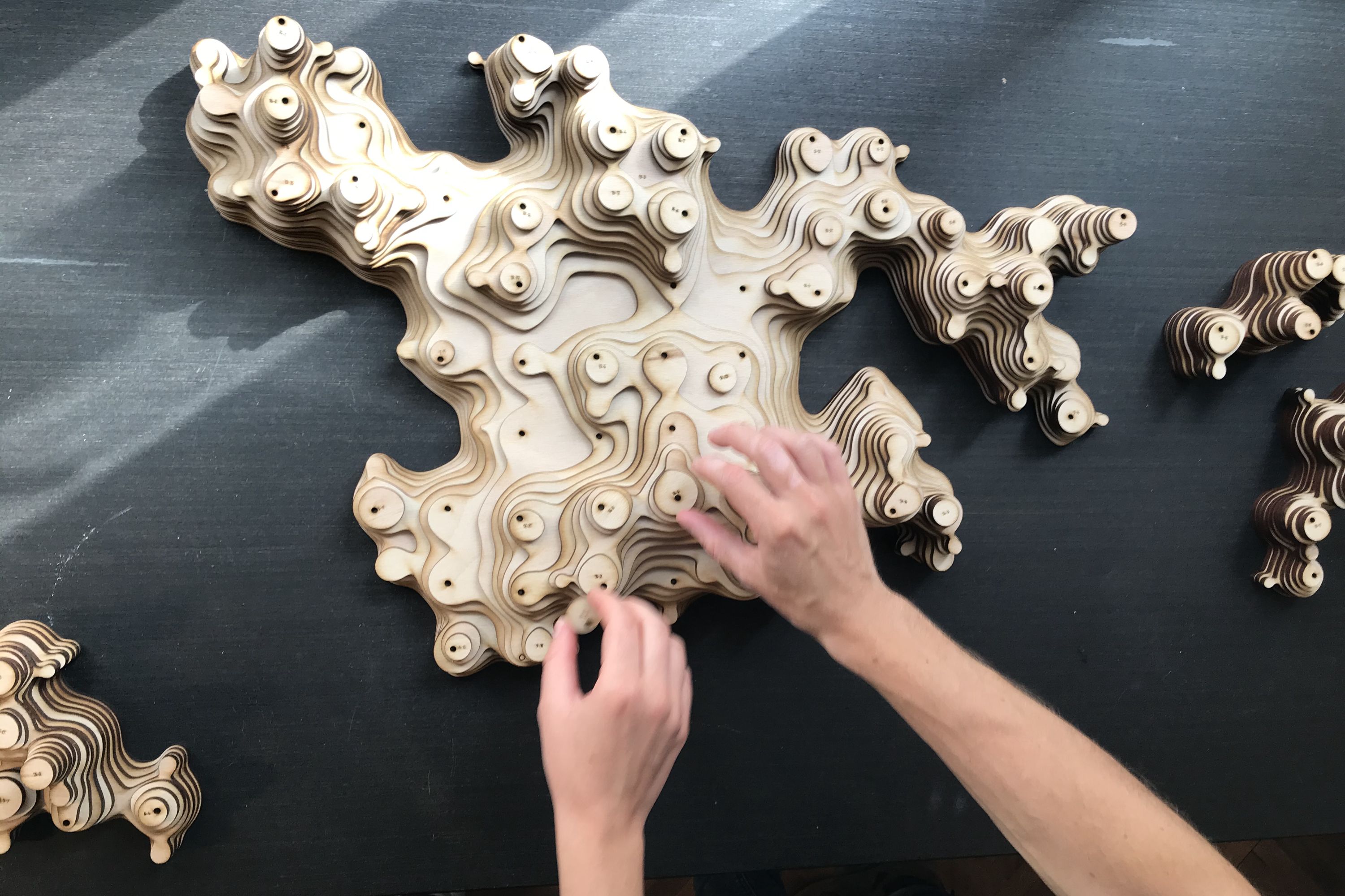 Hands assembling laser-cut plywood modules into a topographic aggregated structure on a dark surface