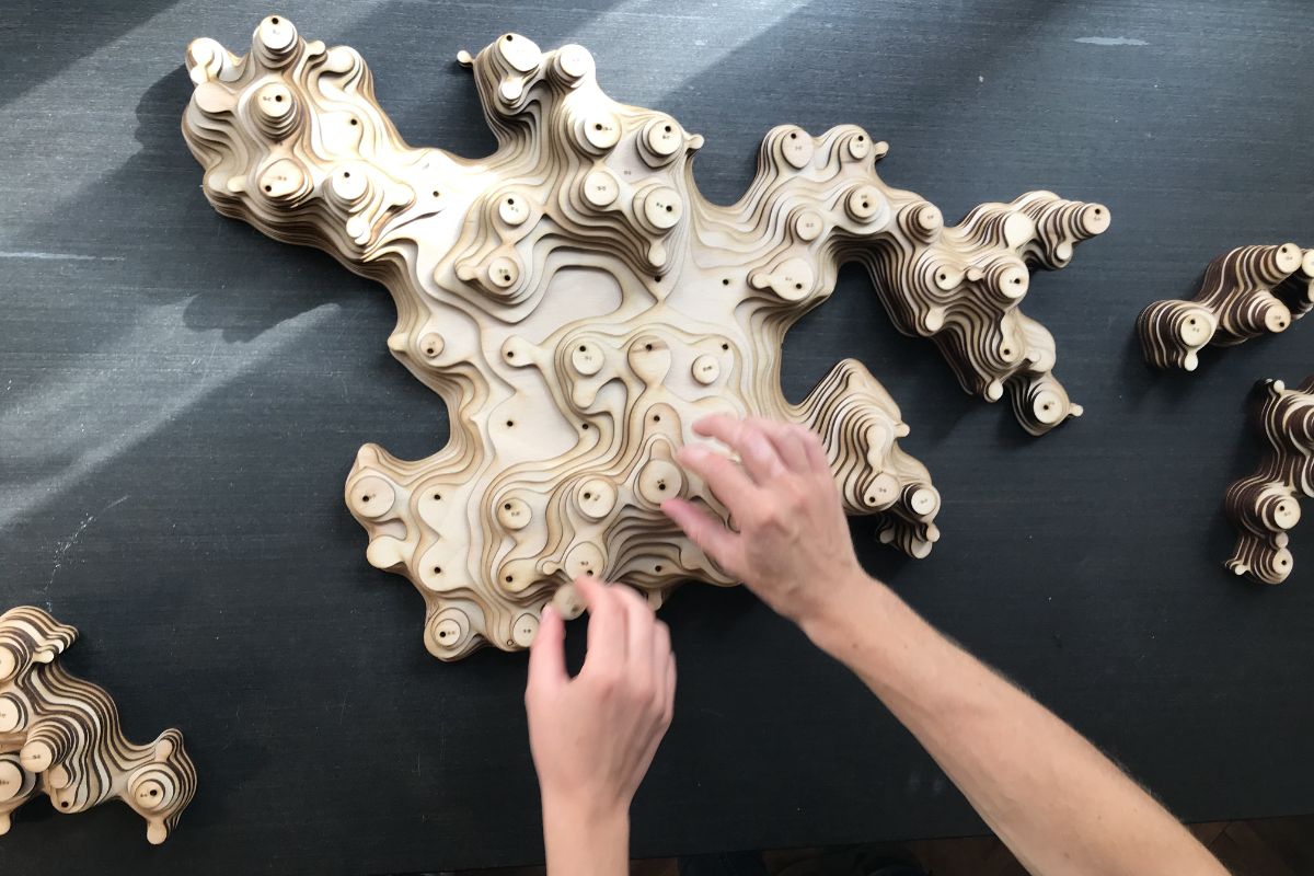 Hands assembling laser-cut plywood modules into a topographic aggregated structure on a dark surface
